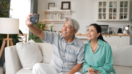 Fototapeta premium Happy grandfather and grandmother making video call with their son at home. Elder man holding smart phone and sitting at sofa while old woman greeting with cheerful vibe at modern house. Myrmidon.