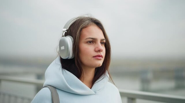Close-up view of lady in casual wear listening to music with headphones, focused expression, blurred background featuring bridge and flowing water, tranquil setting outdoors