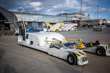 Modern white aircraft tug vehicle parked on an airport runway under a bright blue sky with scattered clouds and terminal buildings in the background