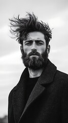Fototapeta premium Powerful portrait of bearded man wearing wool coat with wind blowing through his hair and deep gaze in moody cinematic shot