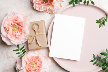Blank white greeting card mockup with kraft gift box and peach roses flowers and green leaves on beige background. Beautiful flat lay with copy space for invitation or thank you card