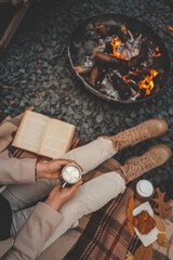 Woman sitting on a blanket by the campfire, holding a metal cup with marshmallows. Warm autumn atmosphere with boots, croissants, book, and cozy outdoor setting