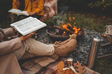 Reading the Bible by a warm campfire during autumn. A cozy scene with boots, blanket, hot drink with marshmallows, croissant, and colorful leaves
