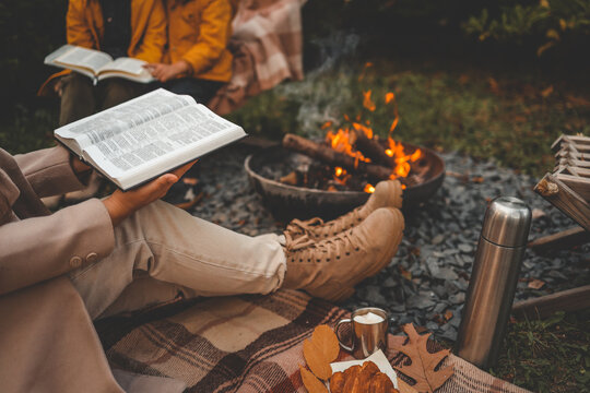 Reading the Bible by a warm campfire during autumn. A cozy scene with boots, blanket, hot drink with marshmallows, croissant, and colorful leaves