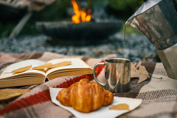 Pouring coffee from a moka pot into a metal cup on a plaid blanket outdoors near a campfire. Cozy autumn morning scene with croissant, book, and fallen leaves