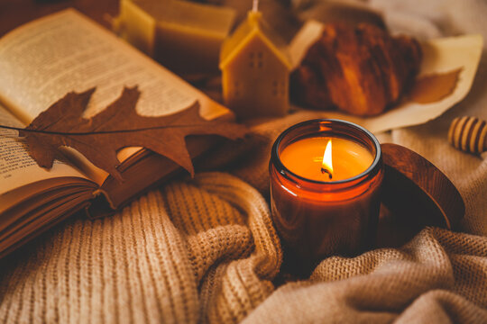Burning amber candle with an open book, autumn leaf, croissant, and cozy knitted textures. Warm hygge atmosphere for relaxing reading time during the fall season
