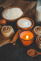 A cozy baking scene with cinnamon buns, a lit candle, flour, sugar, eggs, and milk on a dark surface, perfect for warm, rustic kitchen themes