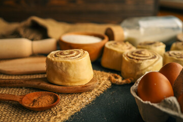 Close-up of raw cinnamon rolls on wooden spoons with baking ingredients like eggs, sugar, milk, flour, and cinnamon on rustic dark background. Homemade pastry preparation