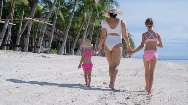 Rear view of young mother enjoys stroling along tropical beach with her two daughters, walking hand in hand, surrounded by palm trees and ocean waves. Slow motion