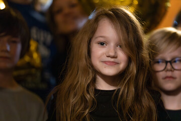 Obraz premium A young girl with long hair smiles at the camera while surrounded by other children at a warmly lit birthday party celebration.