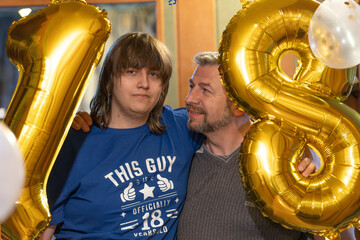 A joyful father and son pose with large golden number 18 balloons, celebrating a milestone birthday in a warmly lit indoor setting.