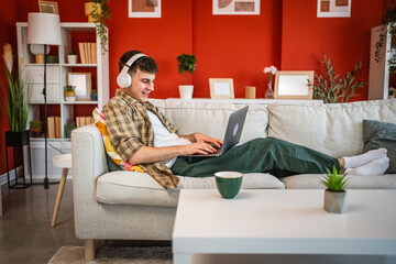 Obraz premium cheerful young man with headphones lying down on sofa and use laptop
