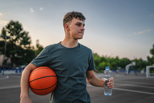 caucasian man teenager open plastic bottle of water outdoor in sunny day drink while hold basketball