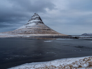 Snowy Kirkjufell mountain and frozen coastal lagoon under dramatic winter skies, Snæfellsnes Peninsula, Iceland. A moody and iconic Nordic scene