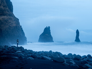 Reynisfjara Beach, Iceland: A lone figure stands against crashing waves, basalt cliffs, and towering Reynisdrangar sea stacks in a moody coastal landscape
