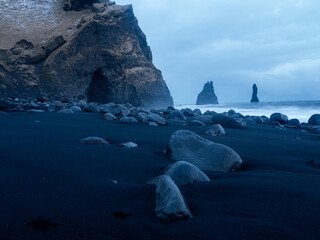 Close-up of smooth volcanic stones scattered on the black sand of Reynisfjara Beach, with basalt cliffs and Reynisdrangar sea stacks emerging from the Atlantic Ocean in the background