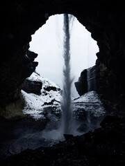 Kvernufoss waterfall as seen from behind, framed by a dark cave opening. The cascading water contrasts starkly with the snow and light outside, creating a dramatic, mystical scene