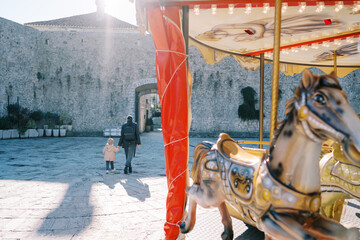 Mother and a little girl walk, holding hands, across the square towards the ancient fortress, past...