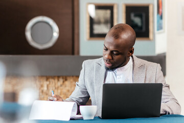 Focused african american businessman working with laptop documents in office holding papers...