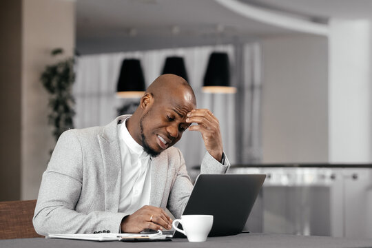 Unhappy young african american male worker in formal suit look at laptop screen shocked by gadget breakdown or operational problems. Frustrated man confused surprised by unexpected error on computer