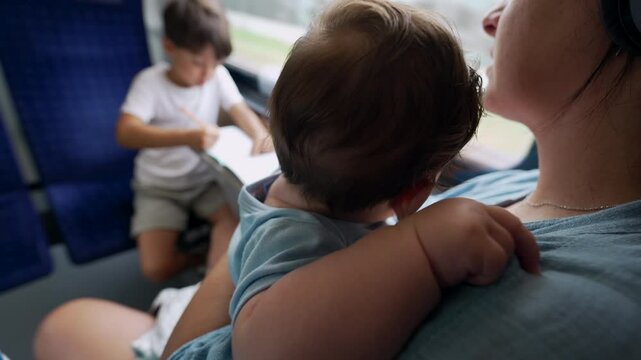 Mother holding baby close to chest on train, older boy drawing in notebook, blue seats, window view of passing landscape, family traveling together on public transport