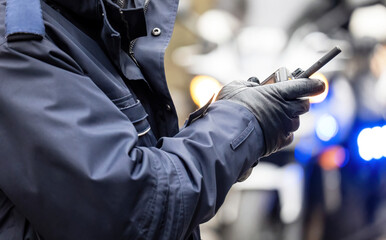 Closeup of a police officer in uniform and gloves holding a two-way radio at emergency scene, blurred crime scene police lights in background, radio communication in public safety services concept