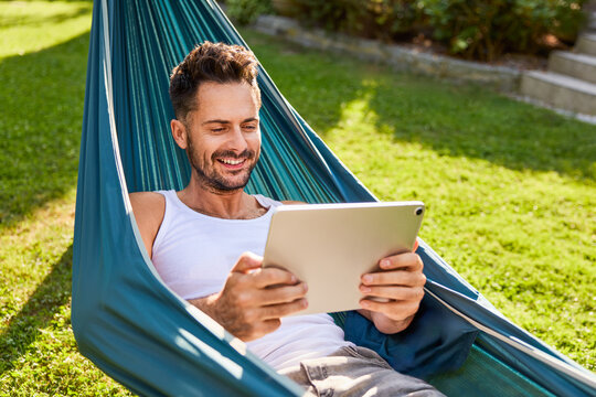 Adult man in hammock using digital tablet relaxing in backyard - Powered by Adobe