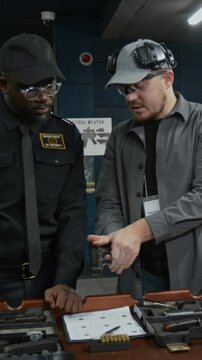 Medium vertical of young professional Caucasian male armorer demonstrating new gun model to African American police officer in uniform at indoor shooting range