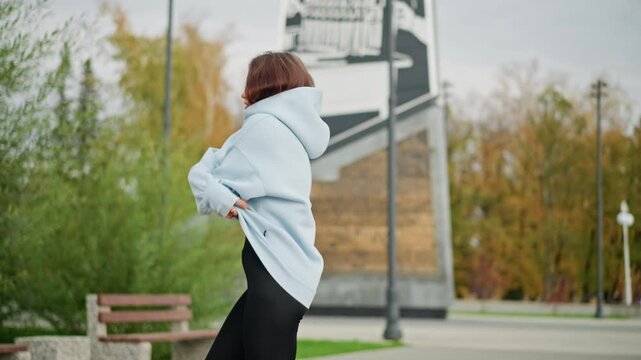 Close-up view of lady removing her blue sweater in park with background featuring buildings, poles, and trees. Ideal for wellness, outdoor, and casual lifestyle moments in serene environment