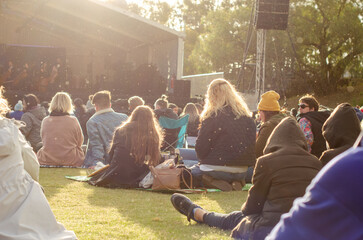A crowd of attendees at an outdoor music festival or concert in a public park, Caucasian people sitting on grass watching a performance on a stage. Warm and relaxed, communal feeling of the gathering.