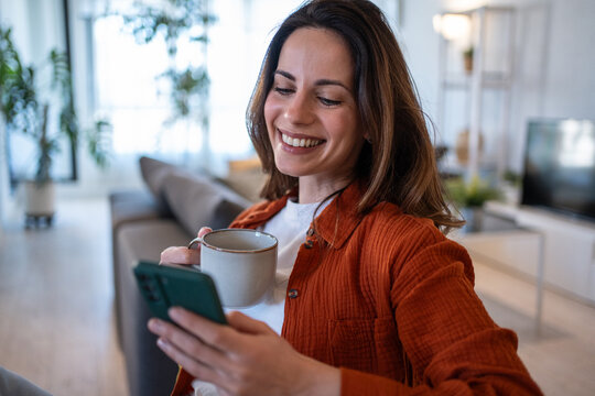 Smiling woman drinks coffee and uses smartphone at home - Powered by Adobe