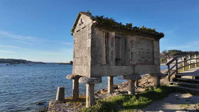 Traditional Galician h&oacute;rreo on a rocky coastline, built from stone with a wooden door and a sloped tiled roof covered in vegetation, overlooking the sea with boats and a coastal village
