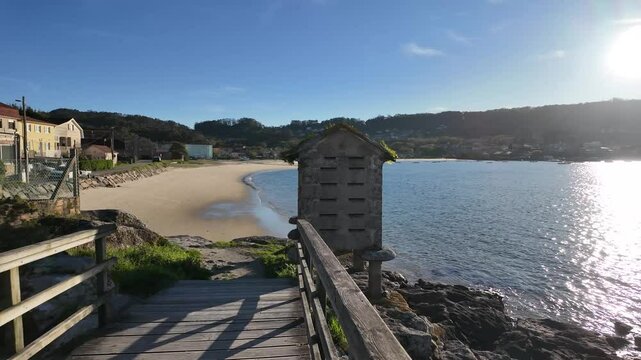 Traditional Galician h&oacute;rreo on a rocky coastline, built from stone with a wooden door and a sloped tiled roof covered in vegetation, overlooking the sea with boats and a coastal village