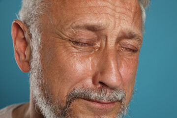 Sad senior man crying on light blue background, closeup