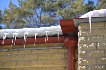 Snow and icicles on the roof of the house against the background of blue sky and pine forest in blur. Drops of water in the air.