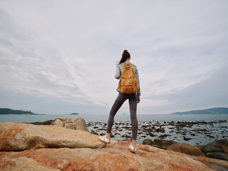 Woman with a yellow backpack standing on rocks, looking at the ocean in a serene outdoor setting, perfect for adventure and travel themes © SHOTPRIME STUDIO