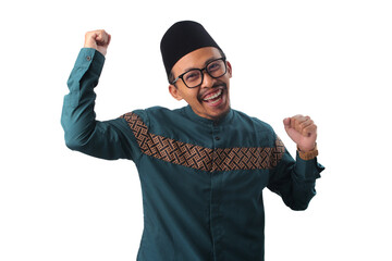 Naklejka premium Excited Asian Muslim man clenching fists in a winner gesture, celebrating success and joy during Eid al-Fitr. Isolated on a white background.