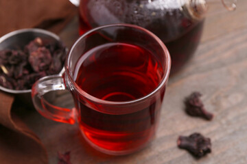 Delicious hibiscus tea in glass cup, teapot and dry roselle sepals on wooden table, closeup