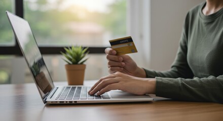 A person using a laptop while holding a credit card, seated at a wooden table near a window with a small potted plant in a well-lit room, focused on online shopping or banking activity.