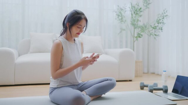 Asian woman resting after home workout and using mobile phone. Young sporty female after practicing yoga, break in doing exercise, relaxing on yoga mat, texting on the cell phone, holding smartphone