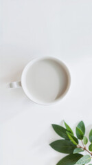 Minimalist composition white ceramic cup, light grey drink, green leaves bottom right, white surface. 