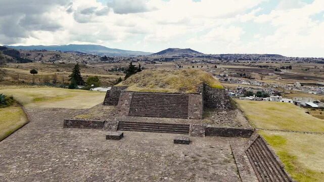 Pyramids of Tlaloc with village and mountains in the background at the archaeological site of Calixtlahuaca in Mexico. Drone camera flies over the pyramids