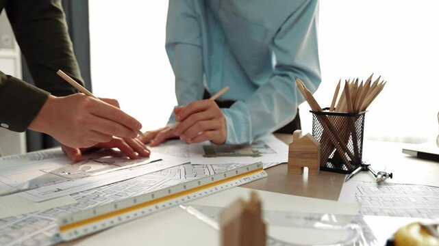 Smart architect and safety engineer discussion about building blueprint on table with architectural document, measurement tools and wooden block scatter around at modern office. Closeup. Delineation.