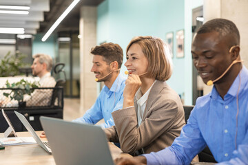 Customer support service. Close-up of caucasian friendly smiling woman, call center operator with headset, working on support hotline in a modern office with a laptop, looking at camera