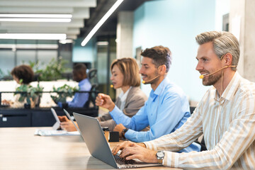 Fototapeta premium Smiling male call-center operator with headphones sitting at modern office with collegues on the background, consulting online.