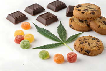 A collection of marijuana-infused edible sweets, including gummy bears, chocolates, and cookies, arranged on a clean white surface. A fresh cannabis leaf is placed beside them, symbolizing medical mar