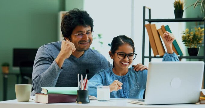 Indian Asian father and young daughter celebrating success together at study table with a laptop, checking results in a modern lavish home, expressing joy, achievement in an educational setting