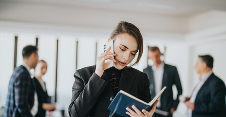 Focused businesswoman multitasks by talking on the phone and checking a planner in a professional...