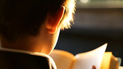 A cinematic slow-motion shot of a young student seen from the back, deeply engaged in reading a book. The setting is a quiet library or a cozy study area with warm, natural lighting.