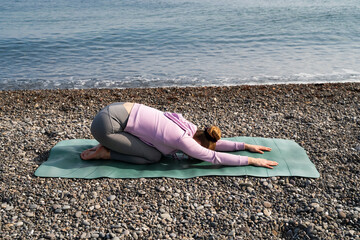 Woman, child's pose on the beach. Sporty girl stretching her back on yoga mat outdoor. Morning relaxing, healthy active lifestyle, fitness, gymnastic, training, warm up, workout in nature, mindfulness